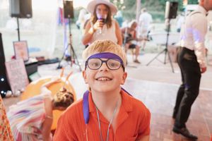 Young boy with glasses wearing an orange shirt smiles brightly at an outdoor party.