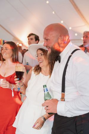 Guests enjoying drinks and laughter at a wedding reception under a white tent with string lights.
