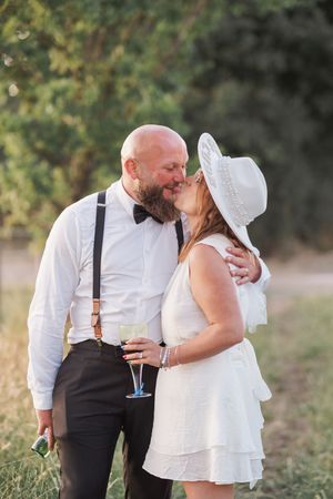 Romantic couple sharing a moment outdoors, with groom in suspenders and bride in white dress and sun hat.