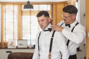 Two people in formal attire making final preparations in a sunlit room with wooden blinds.