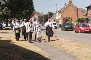 A group of formally dressed people walk along a suburban sidewalk with residential houses in the background.