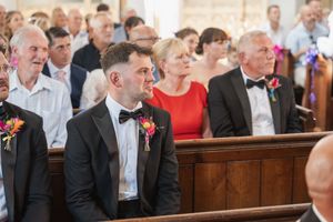 Wedding guests seated in wooden church pews watching the ceremony with attentive expressions.