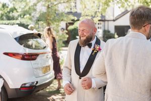 Elegant wedding guest in white suit and black bow tie stands outdoors next to a parked car.