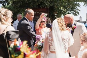 An outdoor wedding ceremony takes place under trees with guests and participants gathered in summer sunshine.