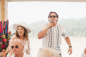Wedding guests enjoy an outdoor celebration under a white tent with colorful decorative streamers in the background.