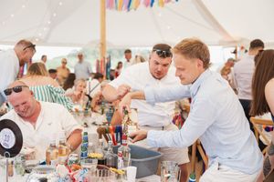 People gather around a table with drinks and beverages under a white party tent with colorful bunting.