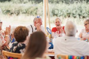 Outdoor wedding reception features guests seated at long tables under a white canopy with natural scenery.