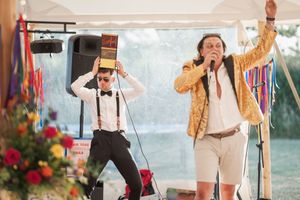 Two performers entertain a crowd under a tent, one speaking into a microphone and another with binoculars.