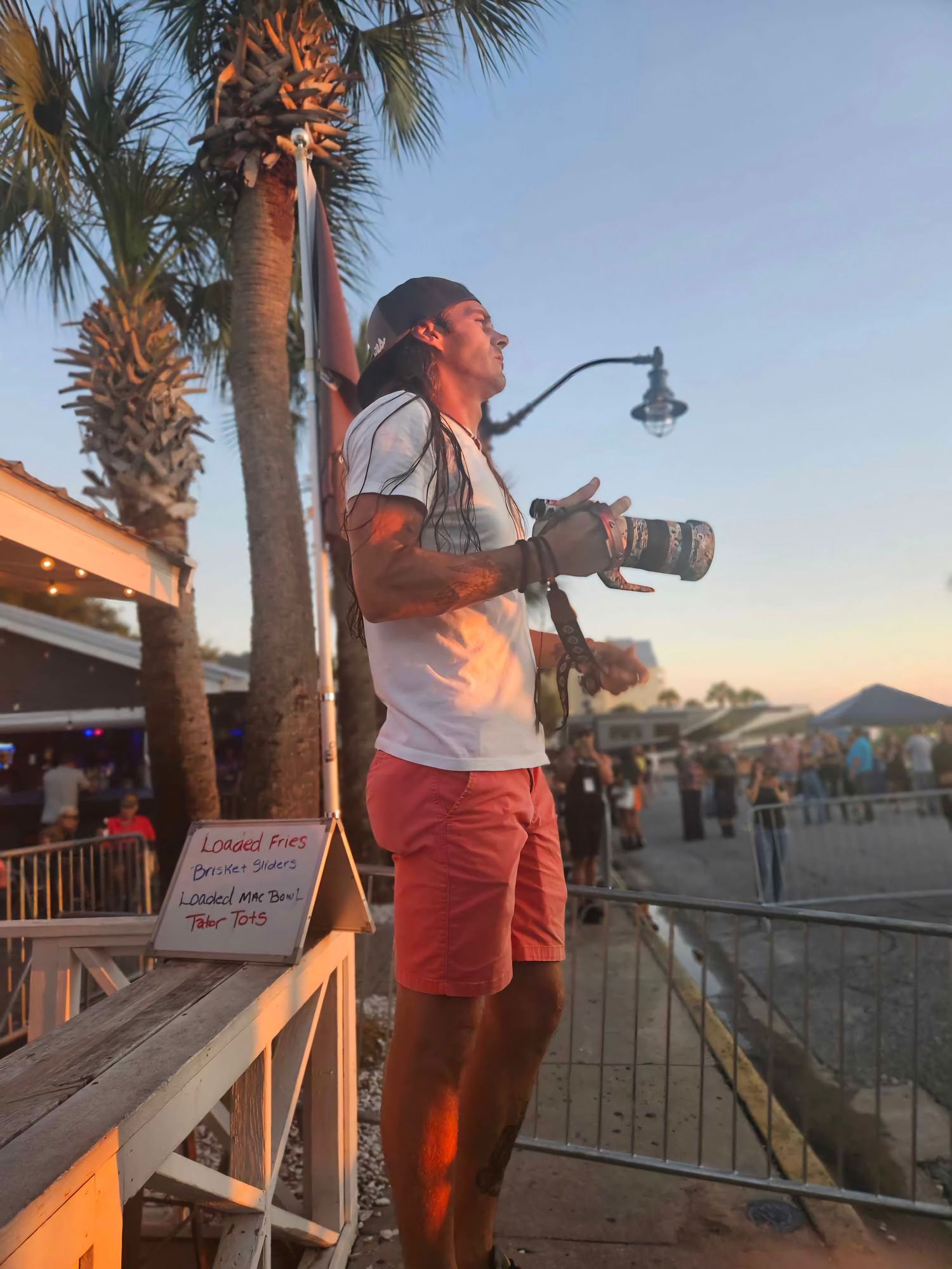 A photographer with a camera stands on a boardwalk at sunset with palm trees and seaside buildings in the background.