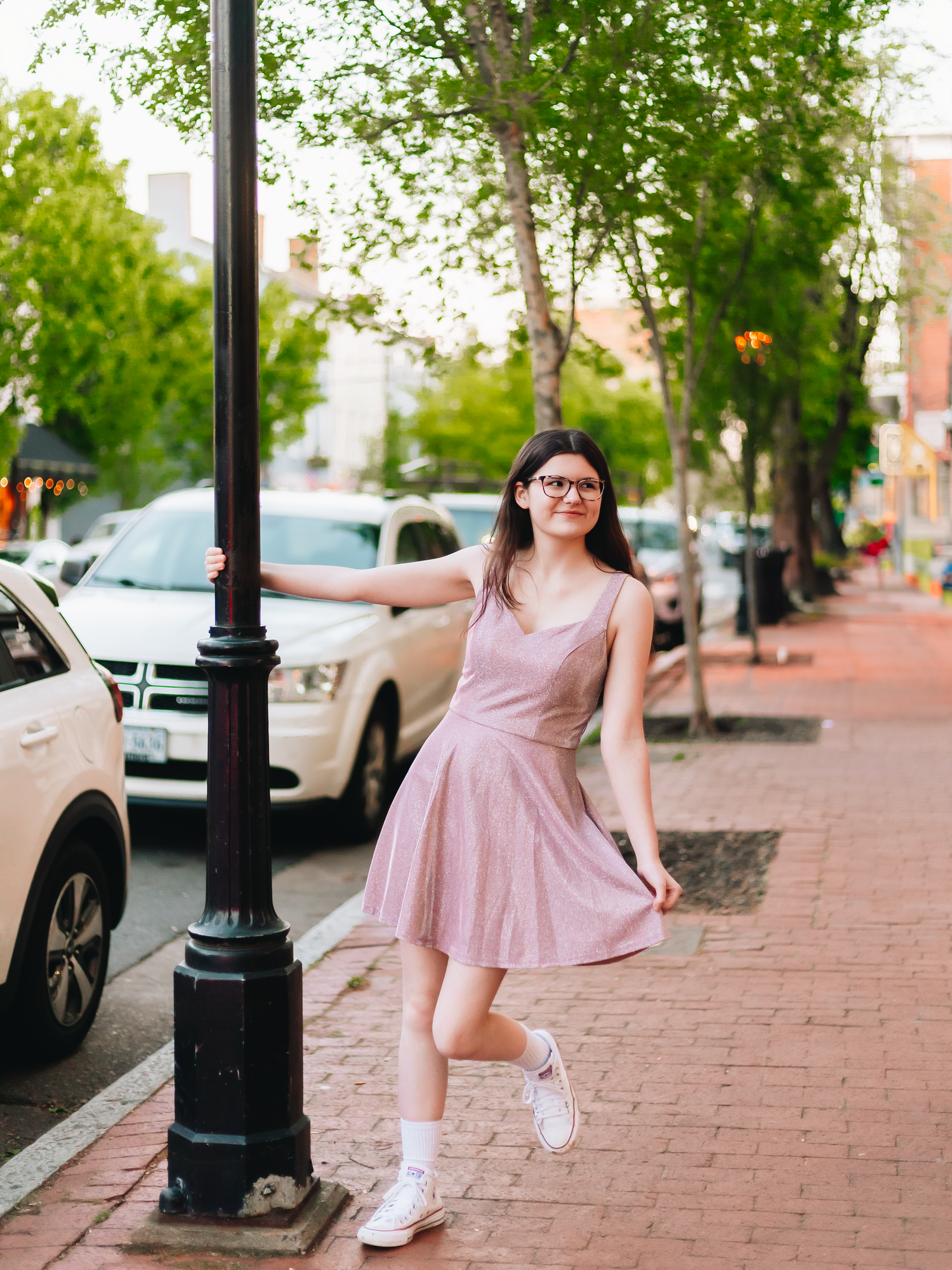 A person in a pink dress twirls playfully on a brick sidewalk lined with lamp posts and parked cars.