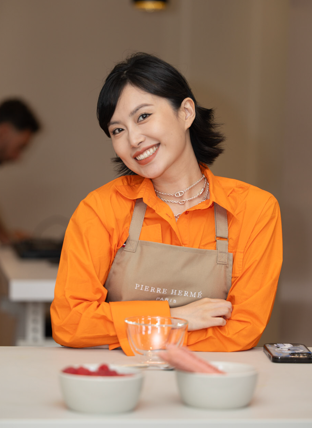 Chef in bright orange uniform and tan apron smiles while preparing food in a kitchen.