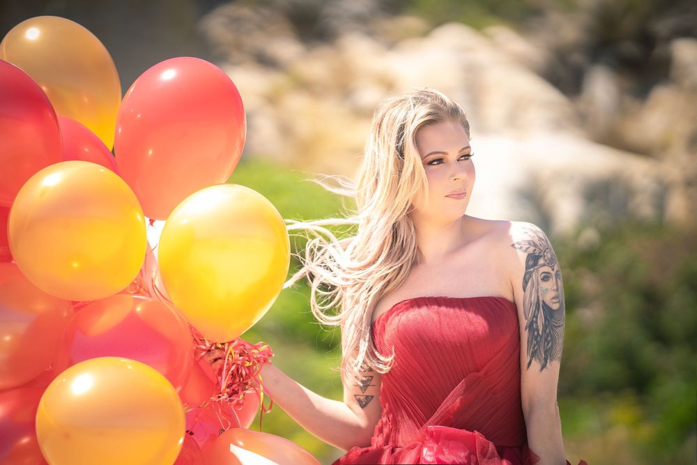 Blonde woman in red dress holding colorful balloons outdoors with flowing hair and tattoo in natural setting.