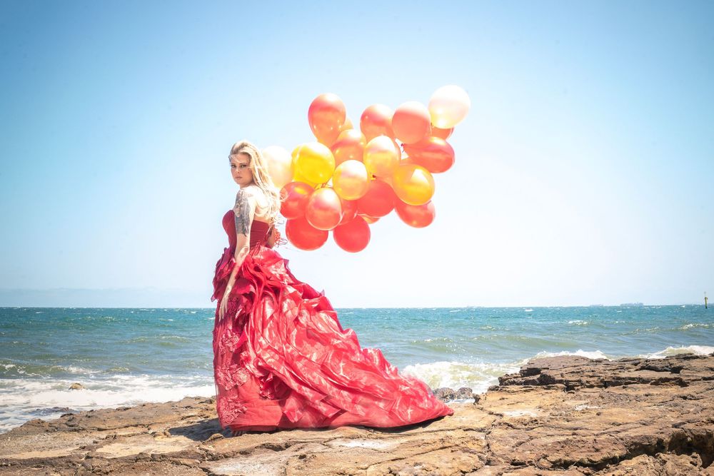 A person in a flowing red evening gown holds orange and red balloons while standing on rocky beach shore.
