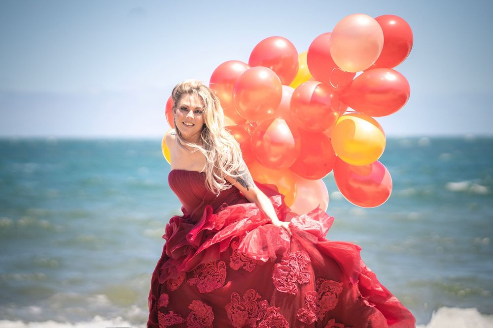 Figure in flowing red gown holds cluster of orange and red balloons on windy beach with ocean backdrop.