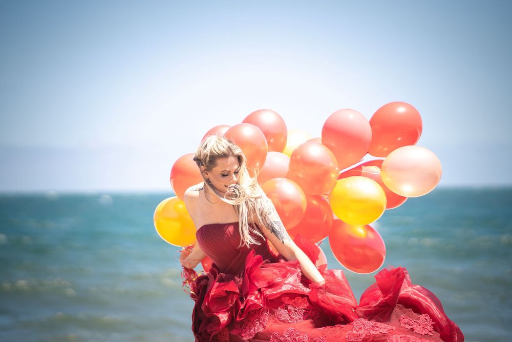 Woman in flowing red dress poses with orange and yellow balloons against ocean backdrop on sunny day.