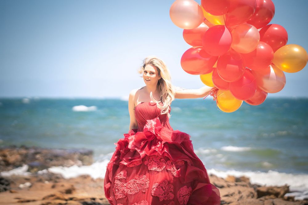 Red and orange balloons float against a blue ocean backdrop on a rocky beach shoreline at sunset.