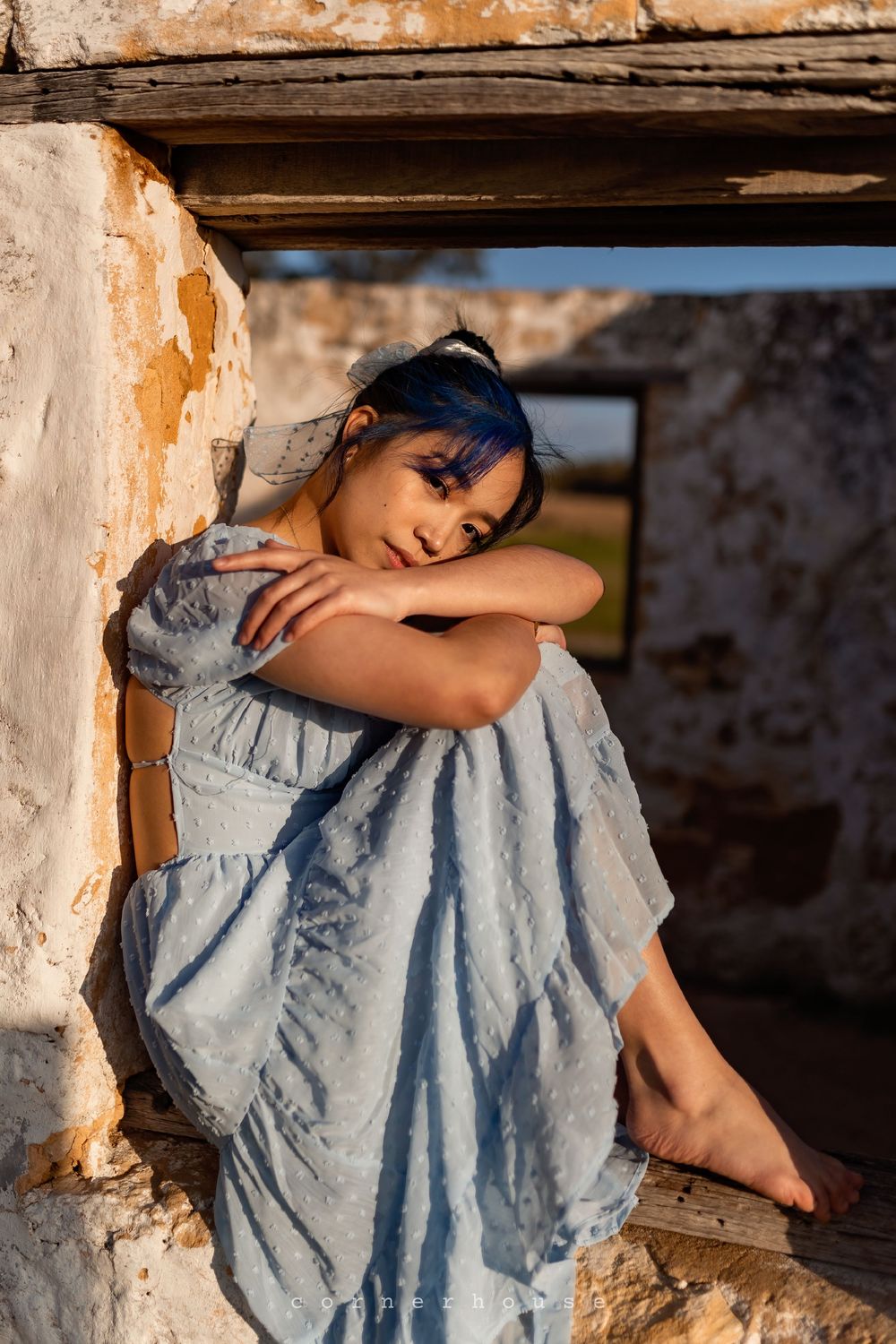 Artistic fashion portrait showing a person in a flowing light blue dress posed against weathered stone walls.
