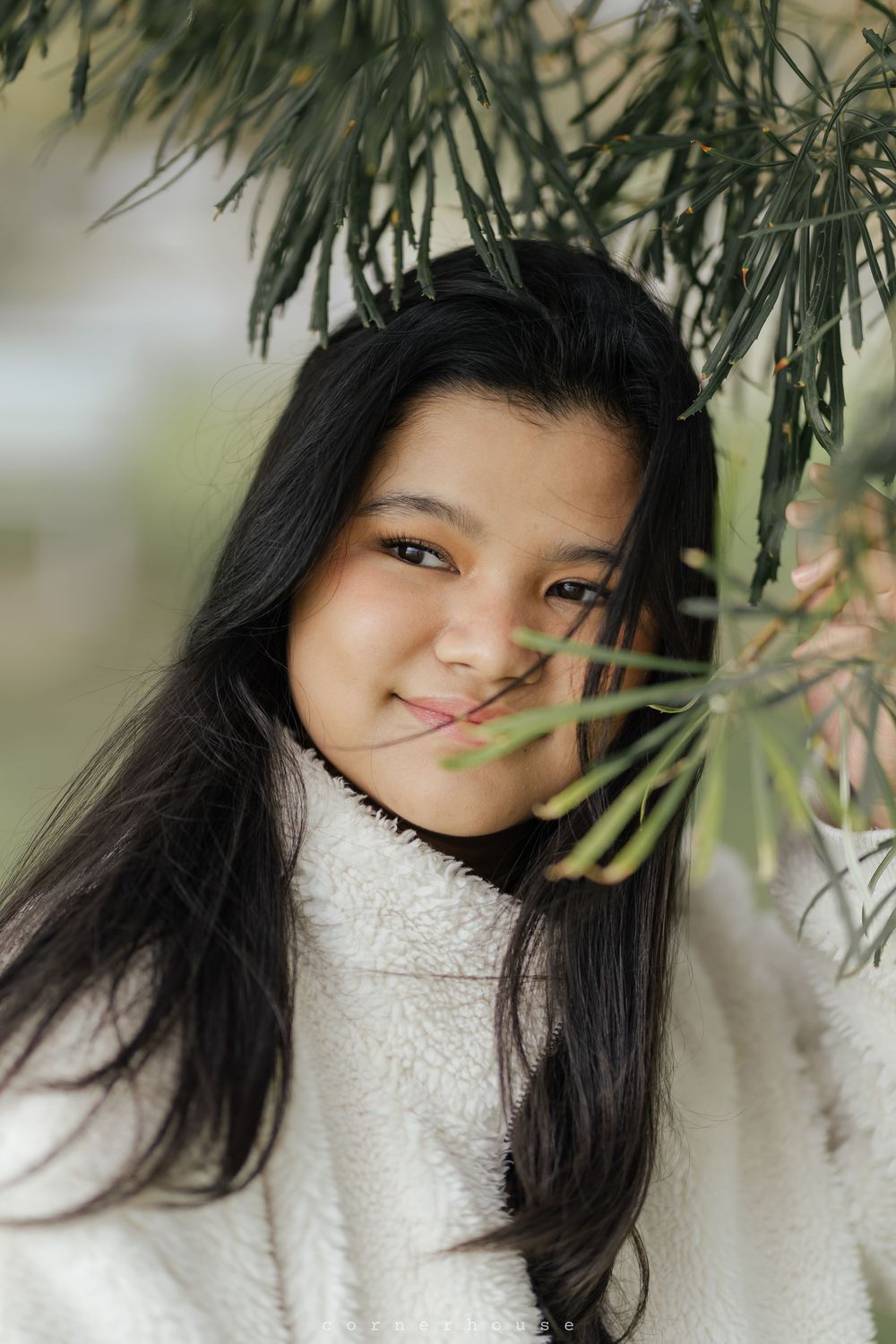Artistic portrait beneath pine branches showing gentle smile and white winter sweater in soft natural lighting.