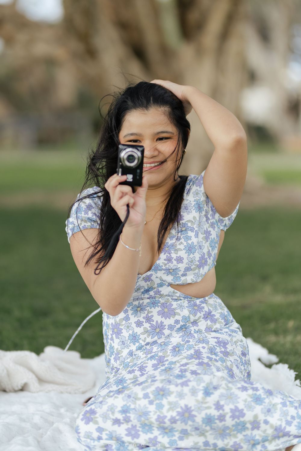 A photographer in a blue and white floral dress lies on white fabric while taking pictures with a vintage camera in a park setting.