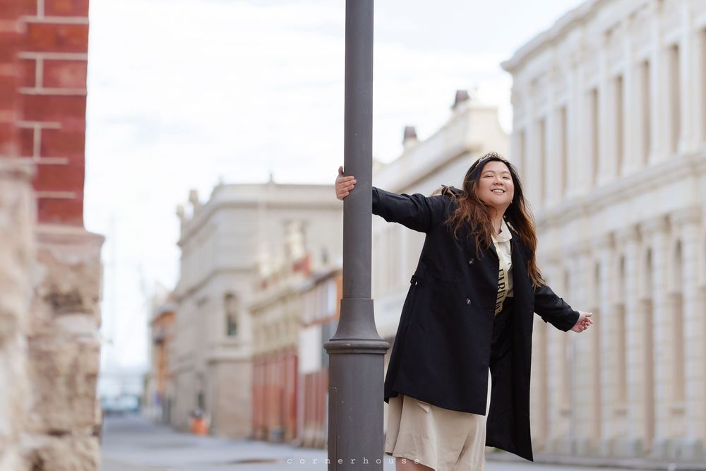 A young woman in a black jacket and light colored dress spins playfully around a lamppost on a European city street.