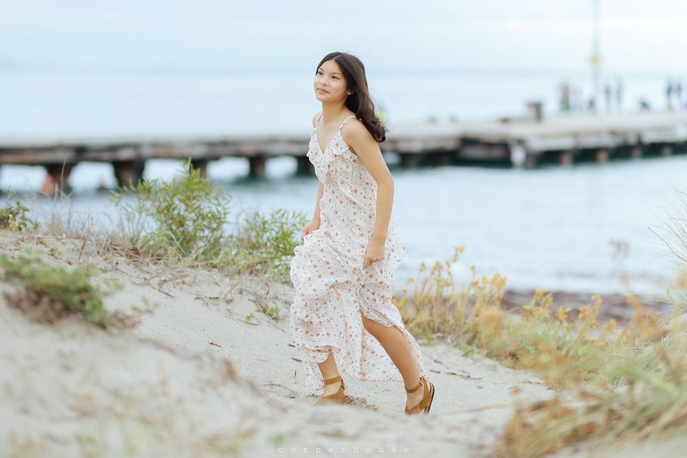 A person in a flowing white dress walks along a sandy beach near a wooden pier on a hazy summer day.