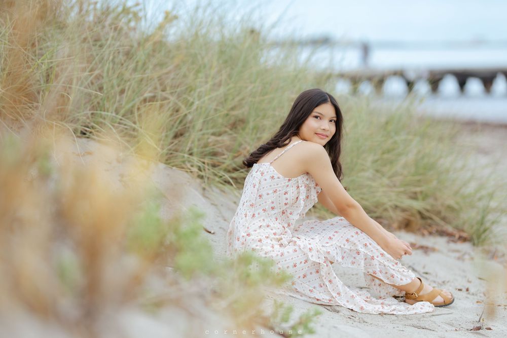 Woman in white floral dress lounges in tall beachside grass during golden hour photoshoot.