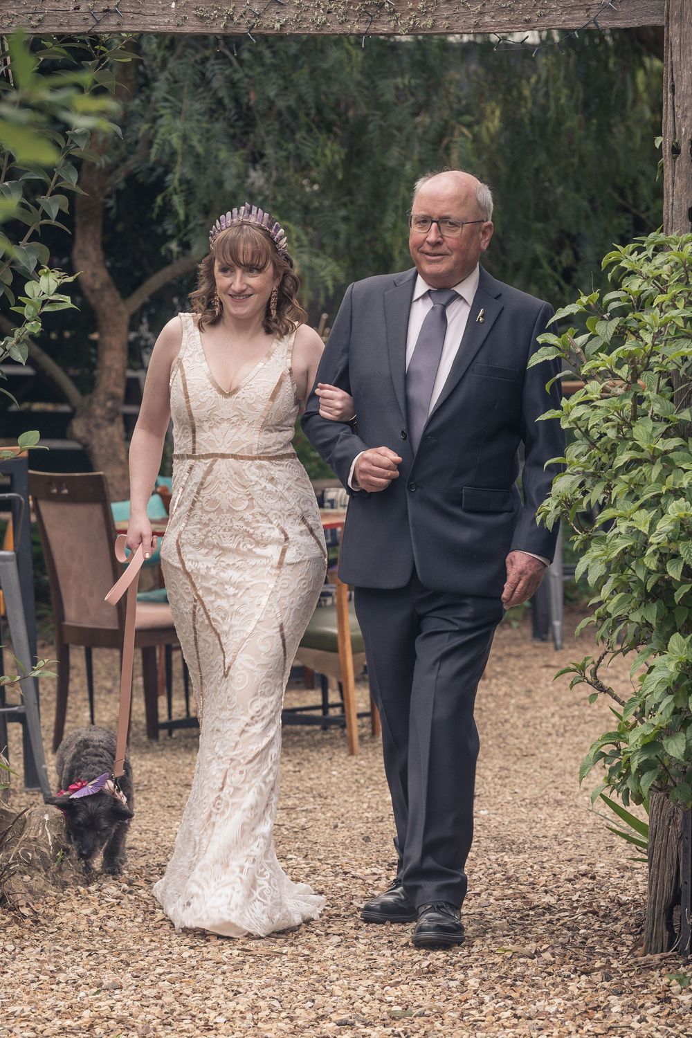 A bride in a fitted lace gown walks down a garden path with her escort while a small dog follows behind them.