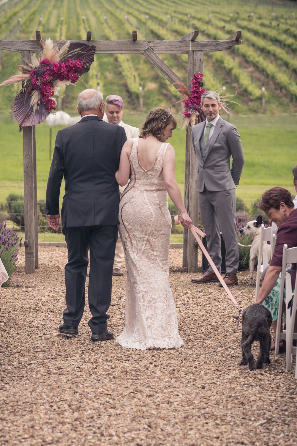 An outdoor vineyard wedding ceremony with attendees gathering under a rustic wooden arch decorated with burgundy flowers.