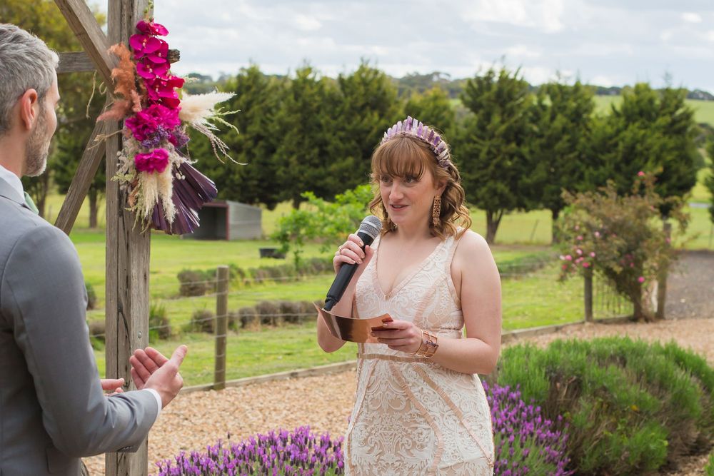 A wedding ceremony takes place outdoors with bright pink flowers decorating a wooden arch and purple lavender in the foreground.