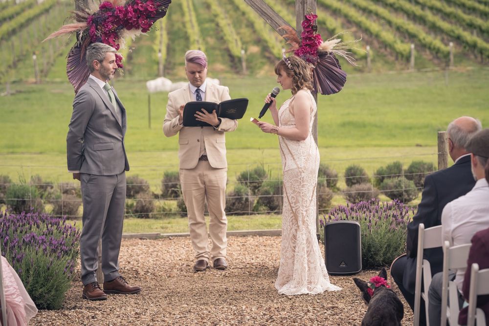 Outdoor vineyard wedding ceremony with people gathered on a gravel path surrounded by lavender and grapevines.