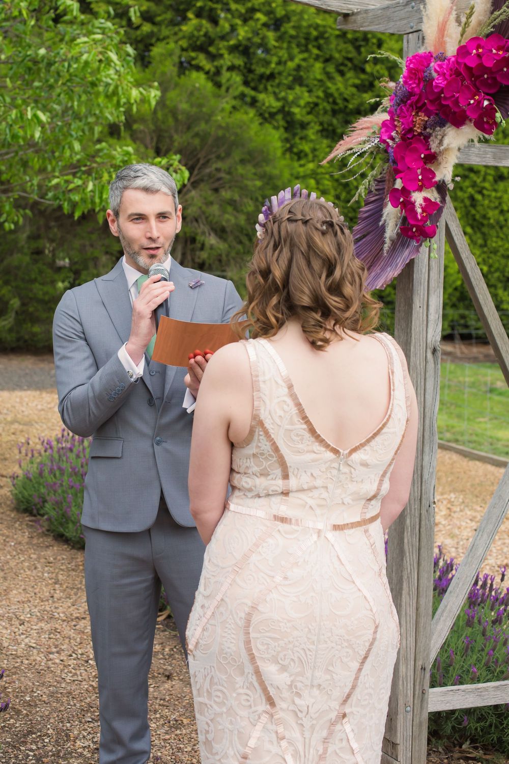 Two people share an emotional moment at an outdoor garden wedding ceremony decorated with vibrant purple flowers.