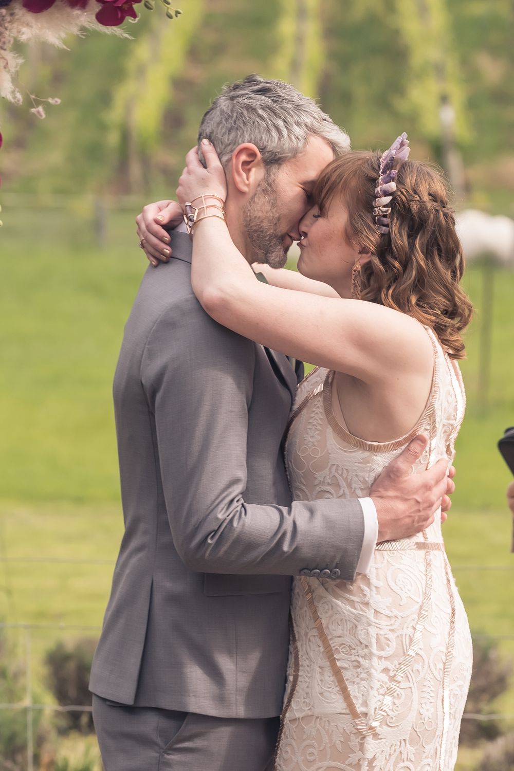 A romantic wedding couple shares an intimate embrace in a garden setting with soft green foliage in the background.