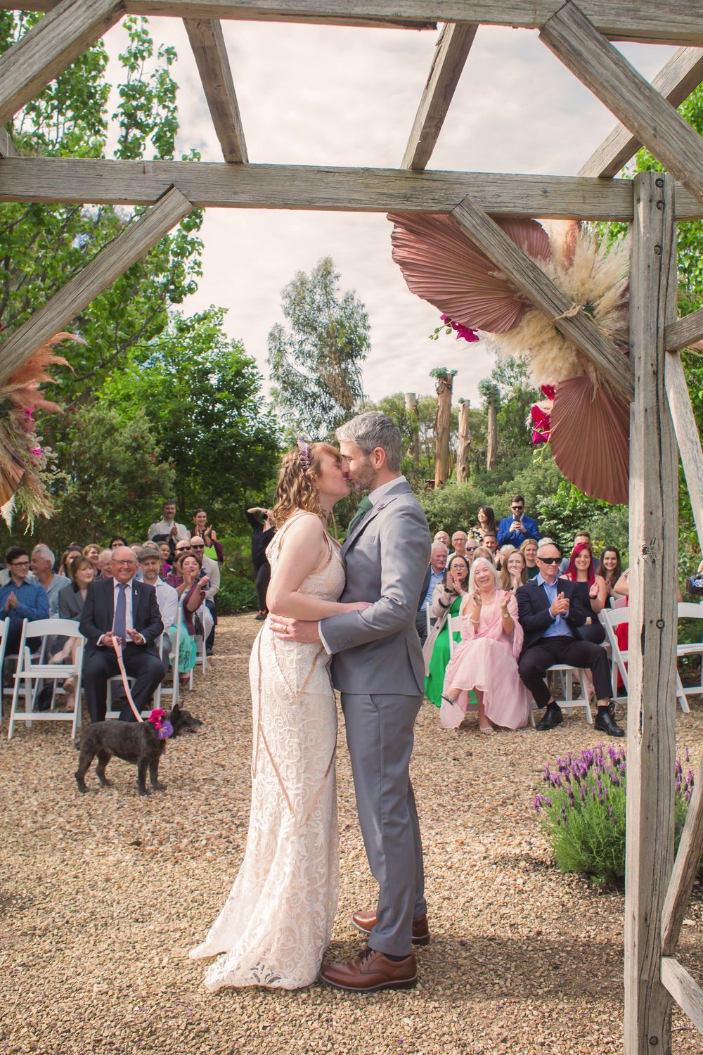 Couple shares romantic first dance under rustic wooden gazebo as wedding guests gather to watch in garden setting.