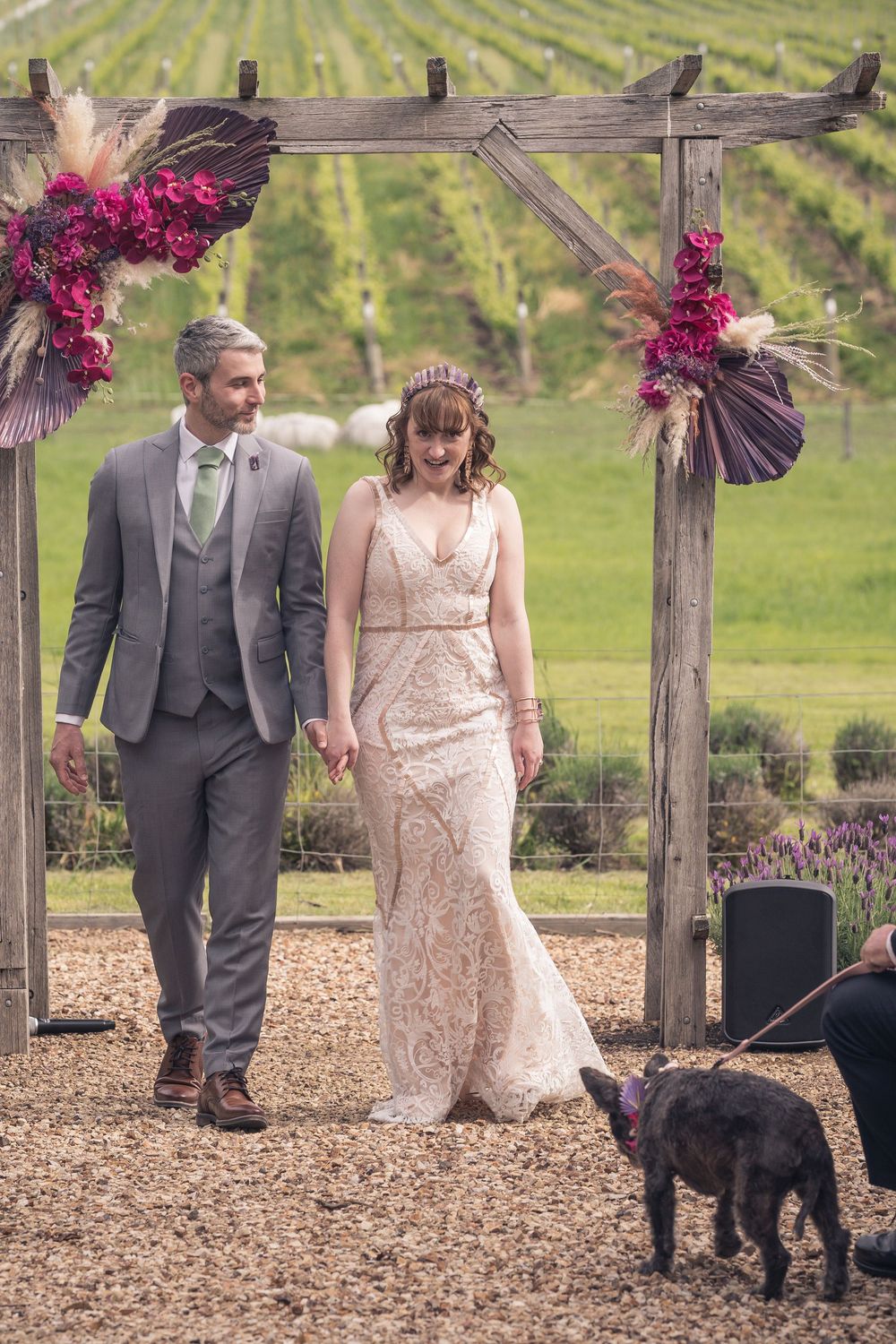 A couple in wedding attire walk hand in hand under a rustic wooden arch decorated with pink flowers at a vineyard ceremony.