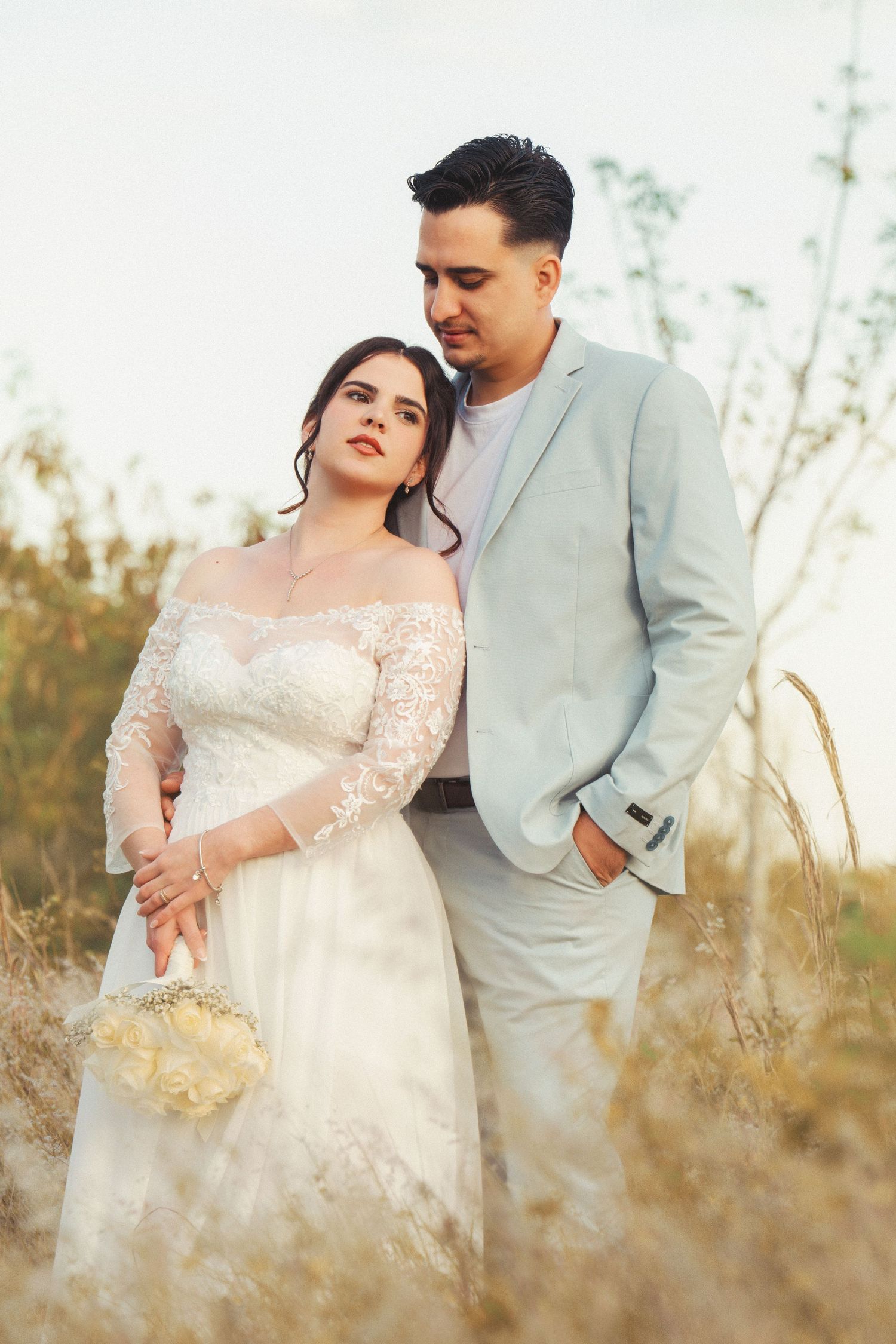 Bride in lace dress and groom in light suit embrace in a sun-lit field during sunset.