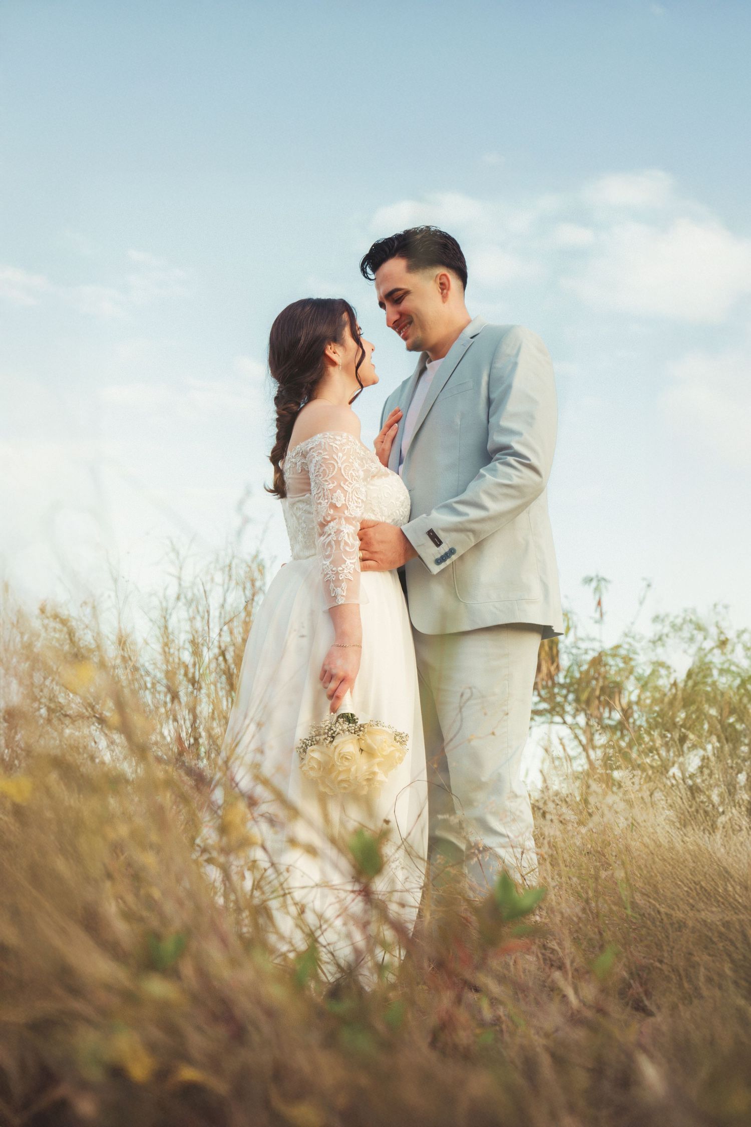 Newlyweds share an intimate moment in a rustic field with blue sky overhead.