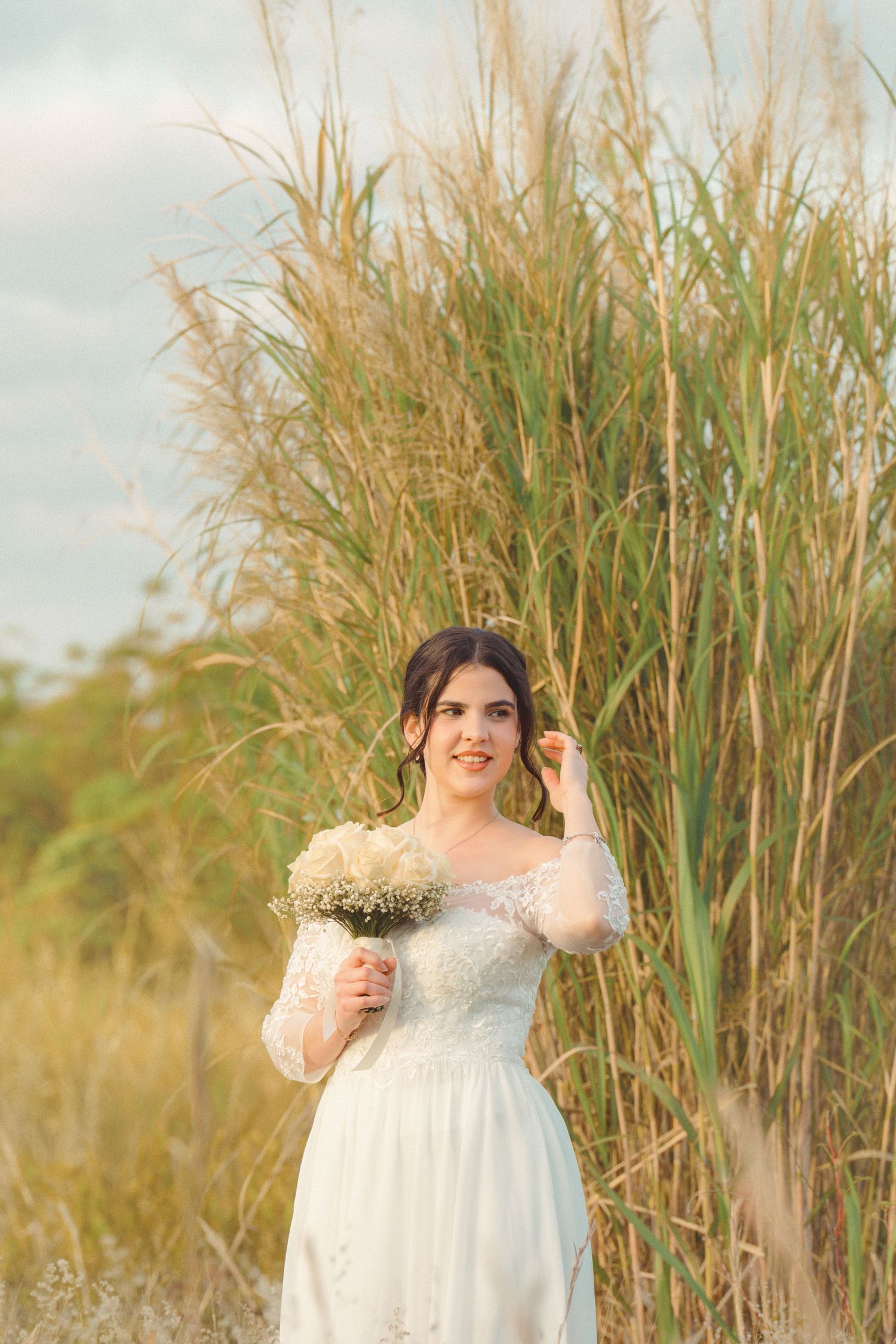 Bride in white lace dress poses elegantly among tall grass in natural sunlight.
