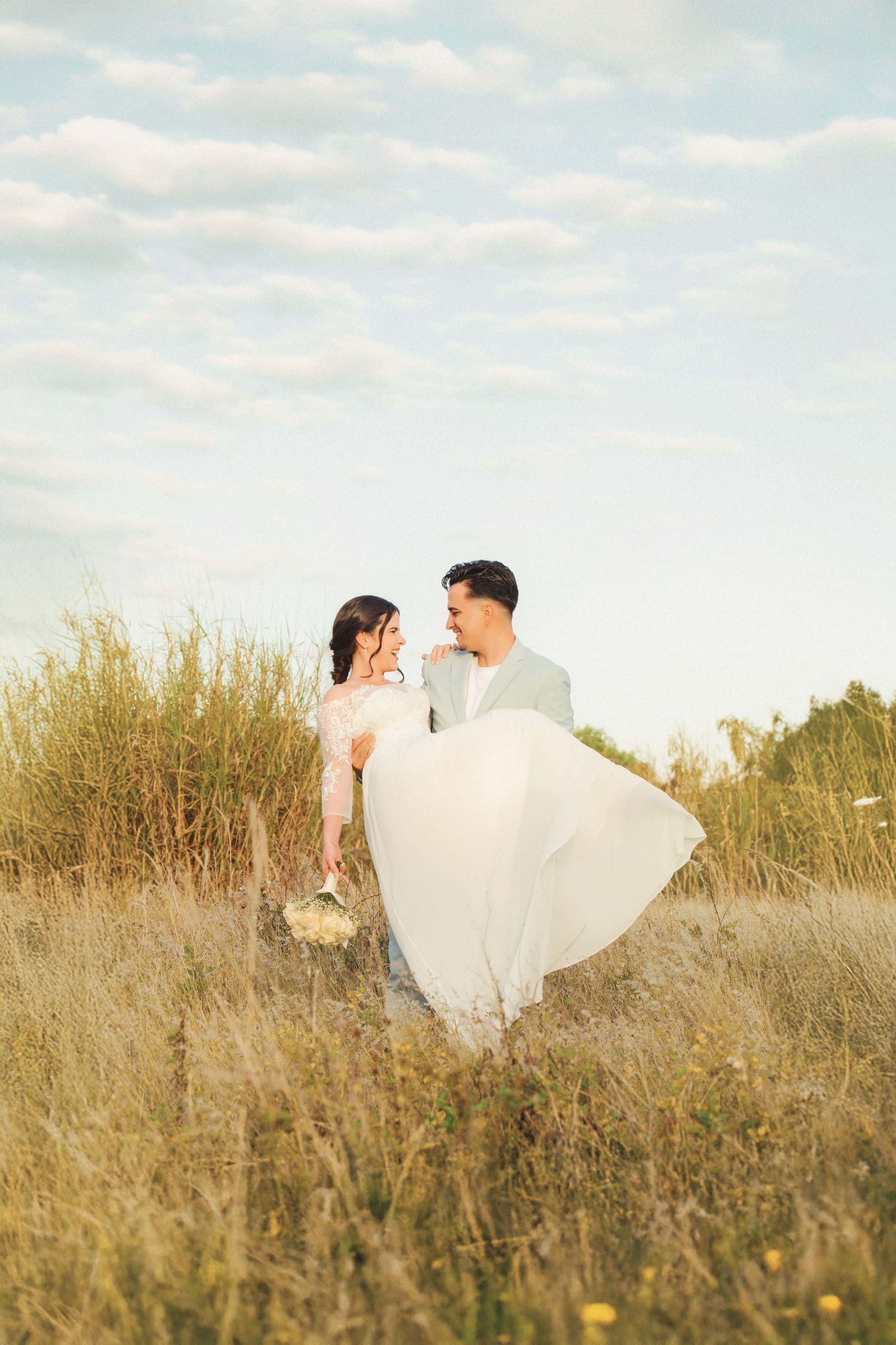 Wedding couple embraces in a field of dried grass under soft clouds.