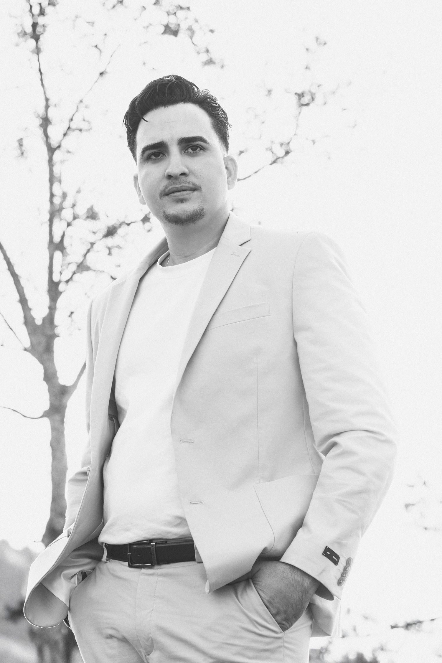 A black and white portrait of a person wearing a light colored blazer and white t-shirt outdoors.
