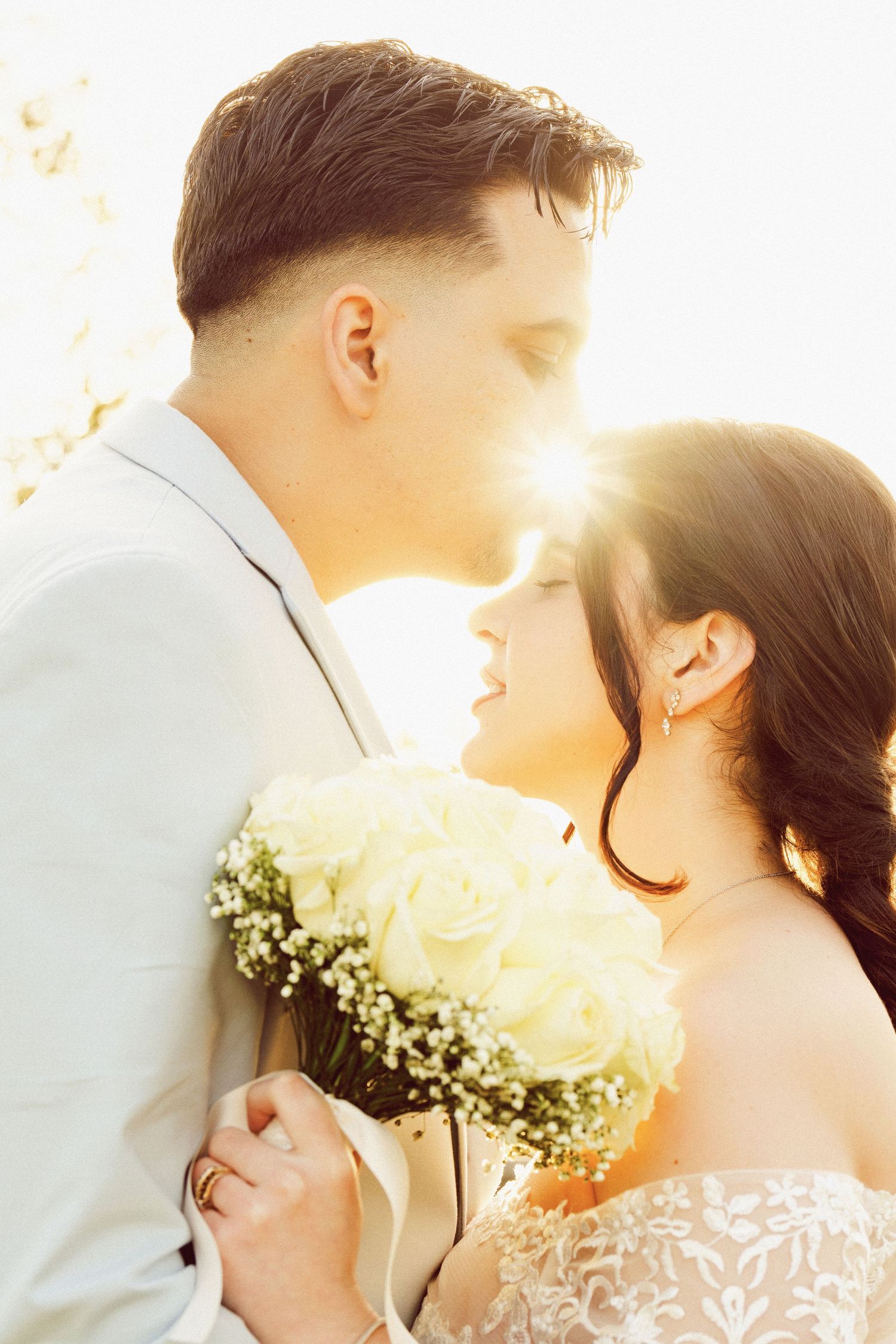 A romantic wedding photo of a couple embracing in golden sunlight with a white rose bouquet.