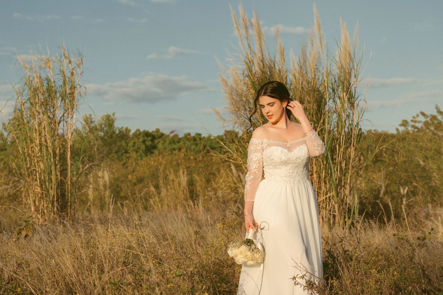 A bride in a flowing white dress stands in a field of tall grass during golden hour.