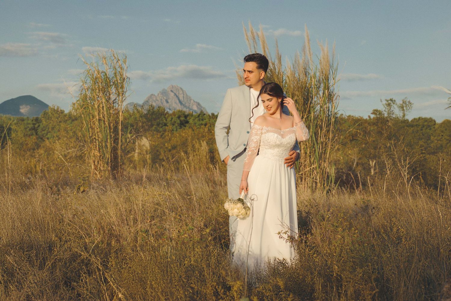 Wedding couple poses in a field during golden hour with mountains in the background.
