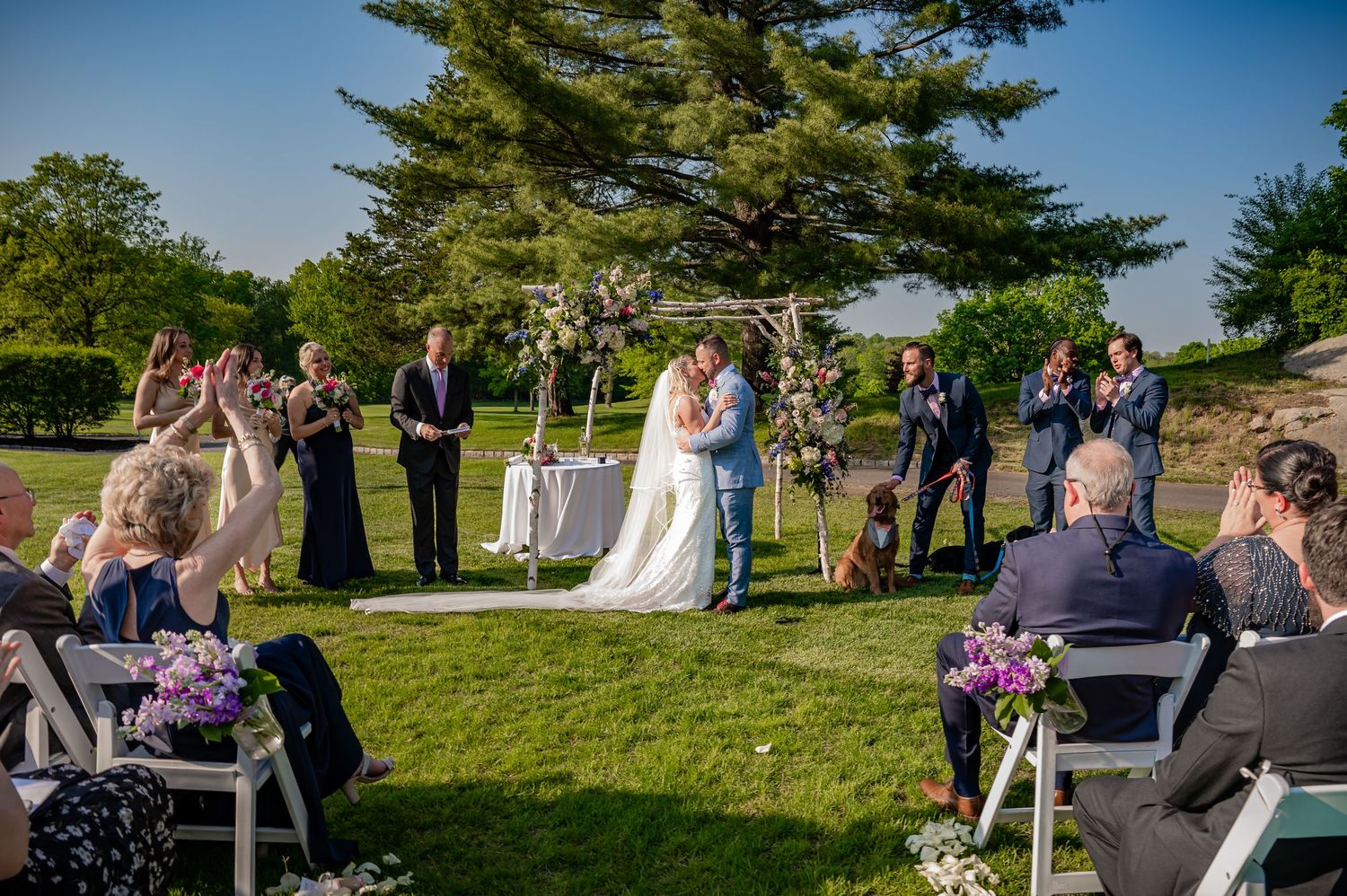 Outdoor wedding ceremony takes place on green lawn with wooden arch and purple floral decorations on sunny summer day.