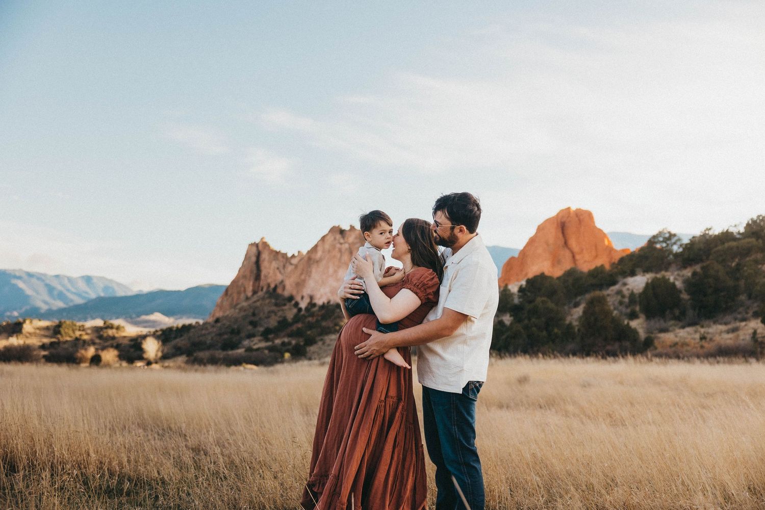 A romantic couple embraces in a golden field with majestic red rock formations and mountains in the background at sunset.