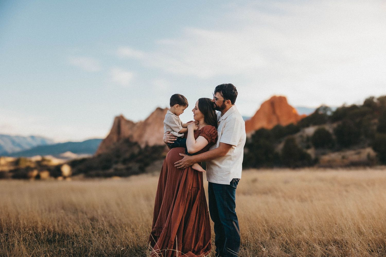 A couple stands together in a golden field at sunset with mountains and red rock formations in the background.