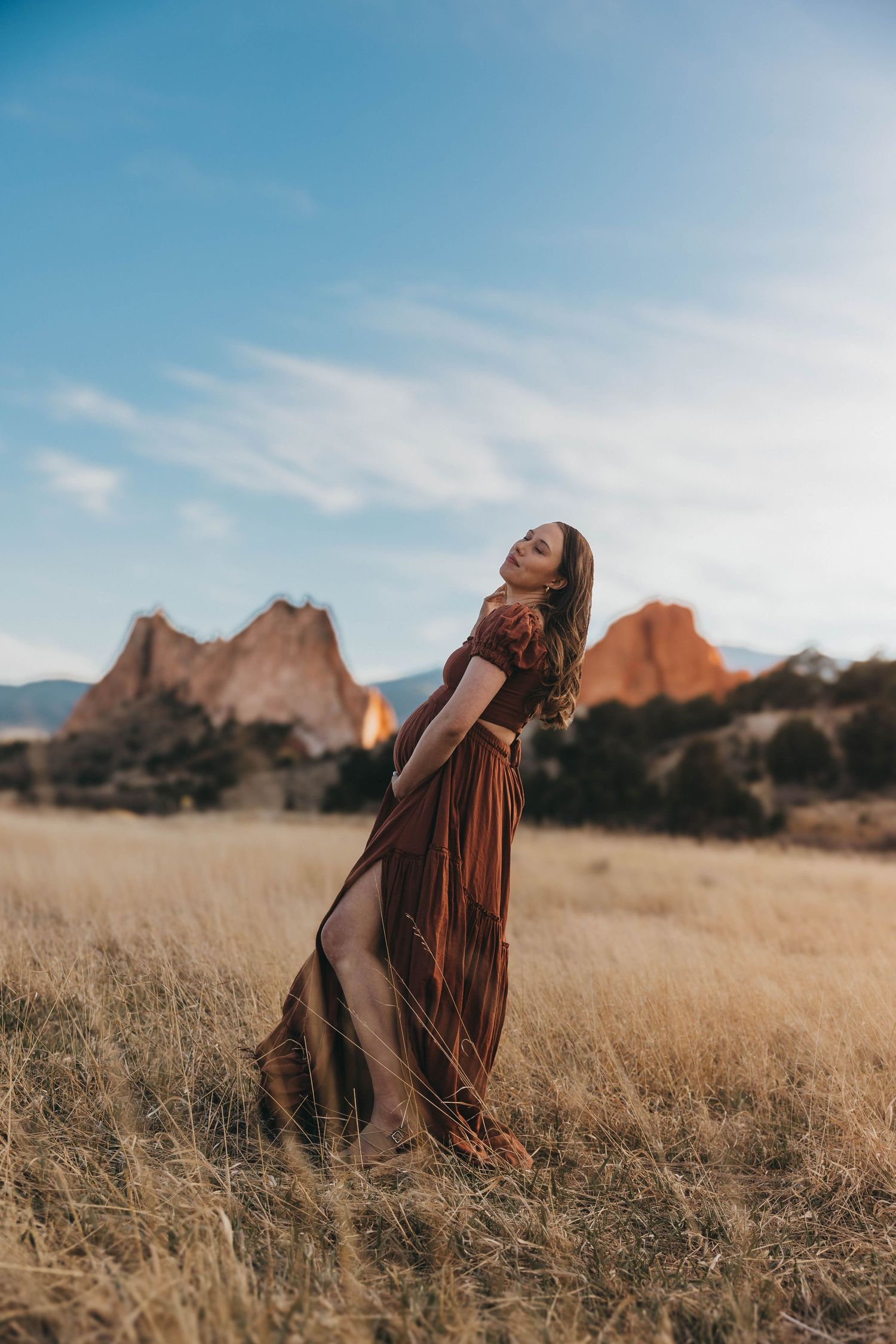 A flowing brown dress catches the wind in a golden field with dramatic red rock formations at Garden of the Gods, Colorado.