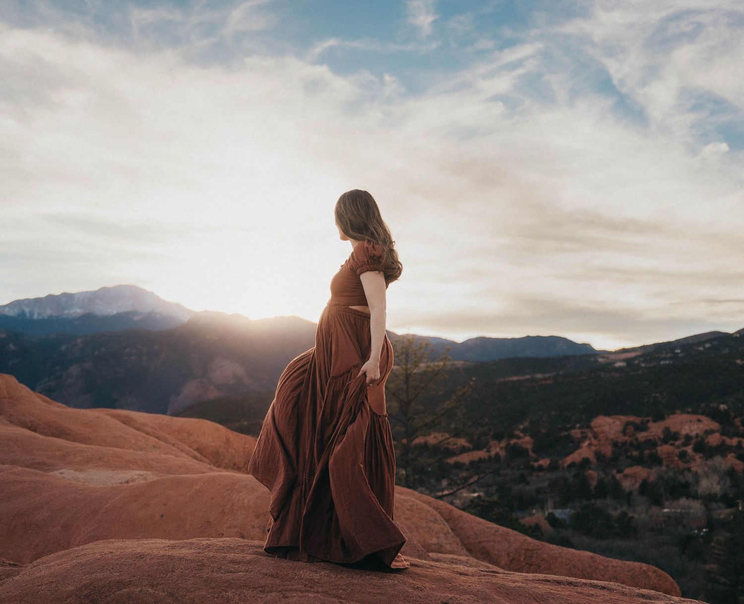 A figure in brown robes sits atop red rock formations at sunset in the desert landscape.