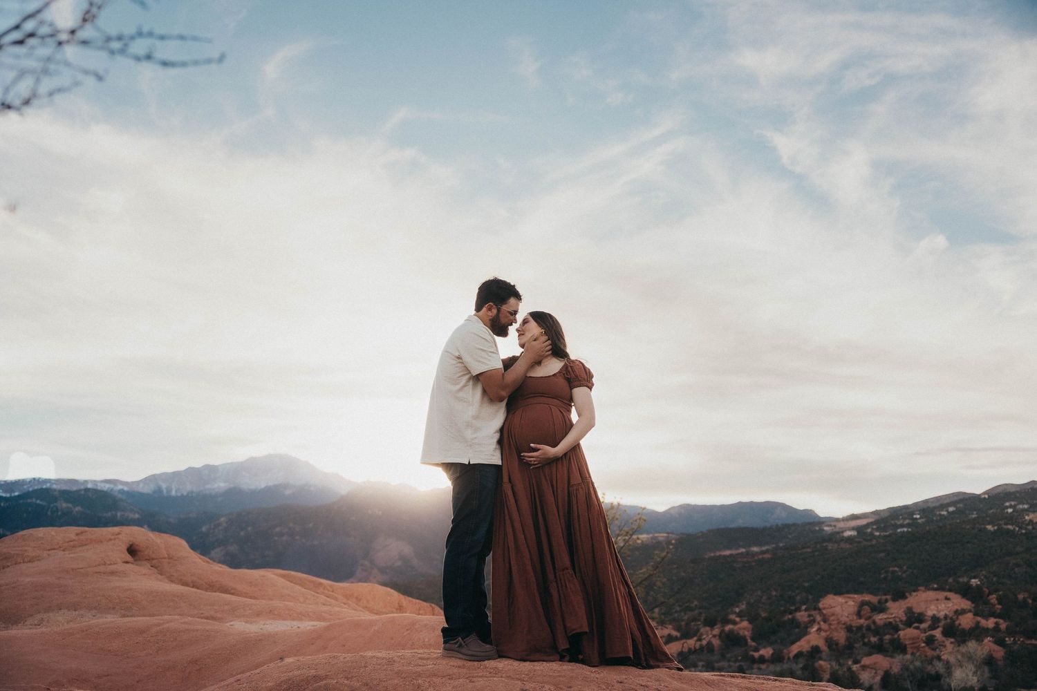 A couple embraces on a mountain ridge at sunset with dramatic clouds and rolling hills in the background.