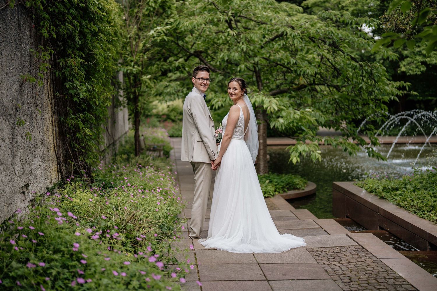 Newlywed couple poses in a lush garden setting with raised flower beds and greenery in the background.
