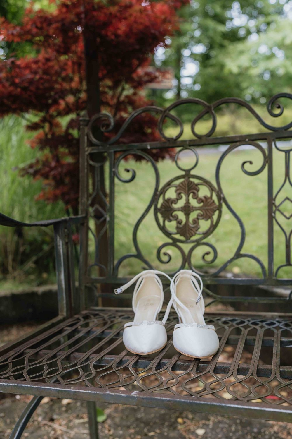 Elegant white bridal shoes resting on an ornate wrought iron garden bench with red maple leaves in background.